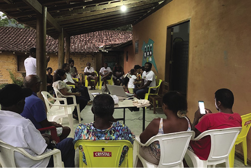 Colour photograph: People sitting in plastic chairs in an open courtyard meeting.