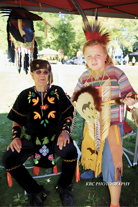 Colour photograph. Two people under a tent canopy. An older man wearing Indigenous regalia is seated on the left, with colourful beaded images and decorations on his black shirt, and a fur hat. There is a large eagle feather fan and other ceremonial items hanging above him. A young boy is standing on the right, wearing a pipebone breastplate, a buckskin apron decorated with an otter, and a feathered headdress. He is holding a fur-trimmed ceremonial item which also has an otter on it.