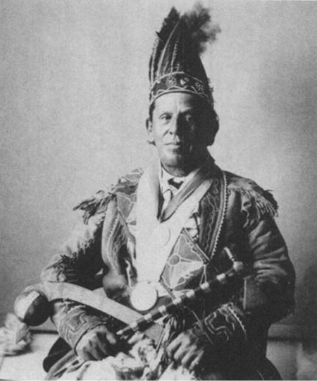 Black and white historical photograph. Portrait of a seated man in Indigenous regalia. He is wearing a feathered headdress, elaborately decorated buckskin jacket, and a large medal on a ribbon around his neck He is holding a tomahawk pipe in his right hand and a club in his left. He faces directly towards the camera.
