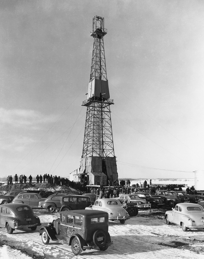 Photo was taken before the well started flowing. It shows a clear day, with snow on the ground. Automobiles and onlookers are gathered haphazardly around the drilling rig. 
