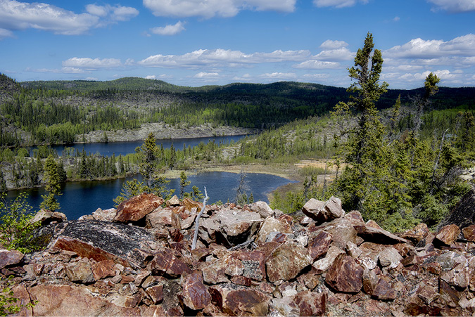 The tailing ponds from gold mines on a bay of Lake Athabasca are nestled by boulders and rocks on hills in the foreground, with forested hills in the background.