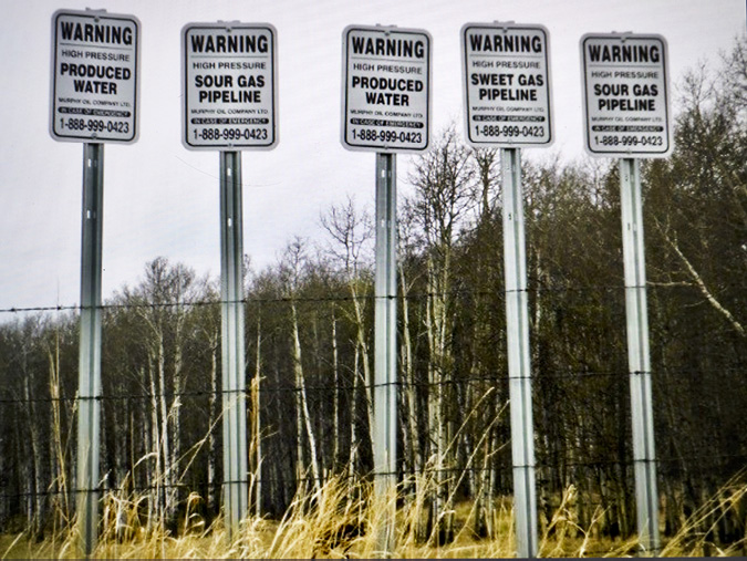 . Warning signs on tall metal posts stand in a row behind a barbed wire fence with dried grasses in the foreground and a mix of aspen and evergreen trees in the background behind the signage. The white signs with black font call out warnings for Produced Water, Sour Gas Pipeline, Produced Water, Sweet Gas Pipeline and Sour Gas Pipelines. Underneath each warning label is a message: In case of emergency call 1-888-999-0423.