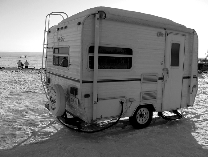 Black and white photograph, a small, white, single-axle caravan sits on a frozen lake, a family poses for a photograph in the background.
