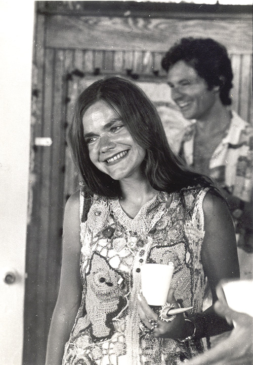 Black and white photo: a woman in a sleeveless smock holding a white cup and wearing a broad smile. A man is in the blur behind her. 