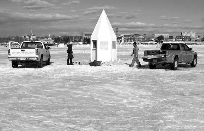 Black and white photograph, a frozen lake with buildings in the background, an octagonal white camera obscura with a conical roof sits in the centre of the image, flanked by a pickup truck on either side, two people in winter clothing approach the structure.