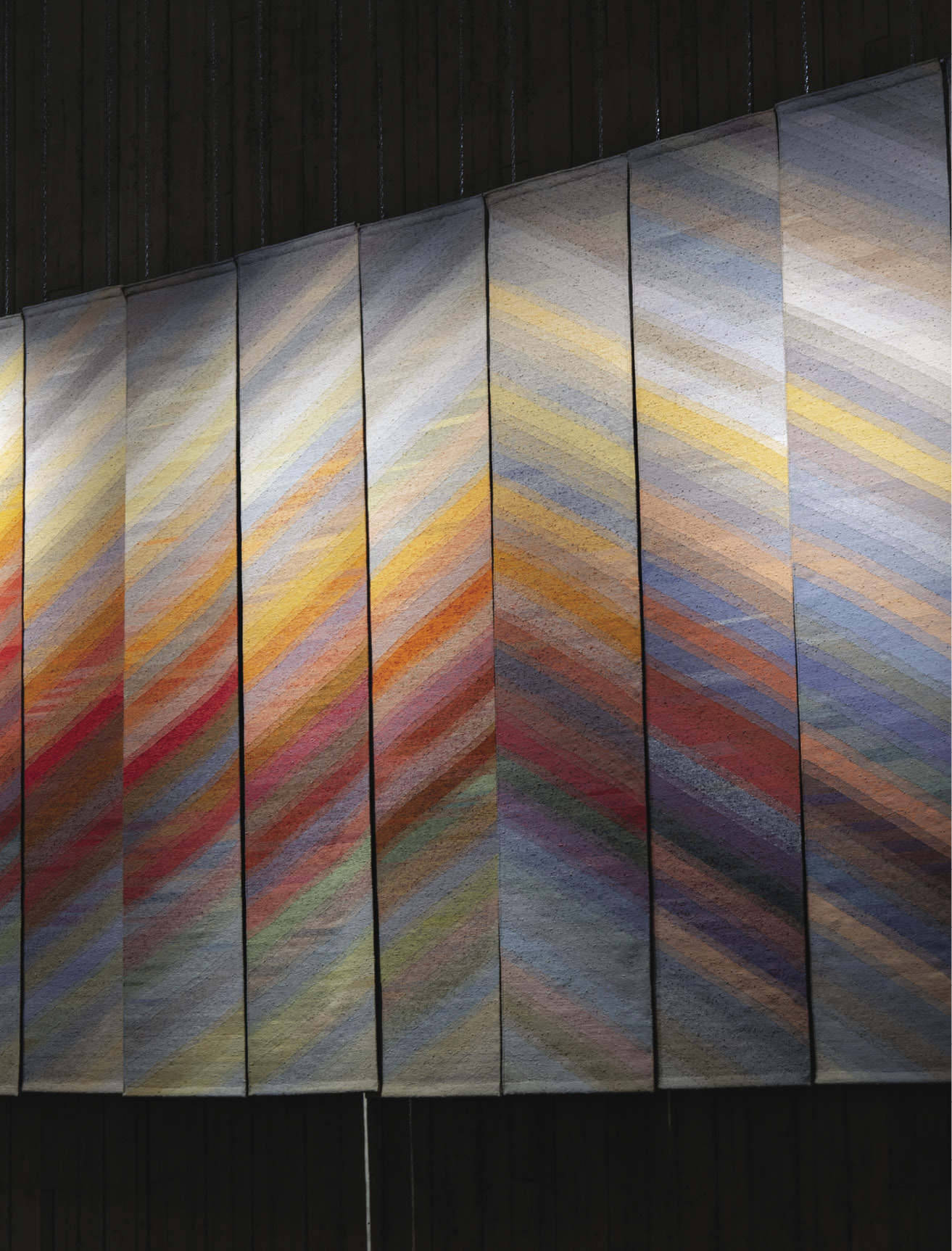 Photo: Twelve tall hangings that from a pyramid pattern from orange, red, pin, and yellow lines against a blue-grey background. 