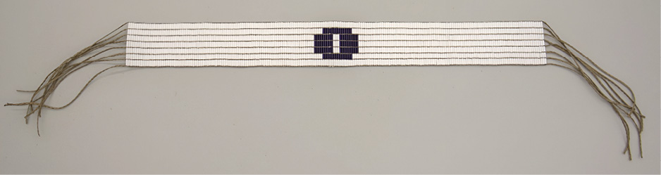 Colour Photograph. Photo of a wampum belt. This is mostly white, with seven rows of wampum. Purple wampum beads, roughly in the shape of an O, take up the centre of the belt. There are leather string fringes at each end of the belt.