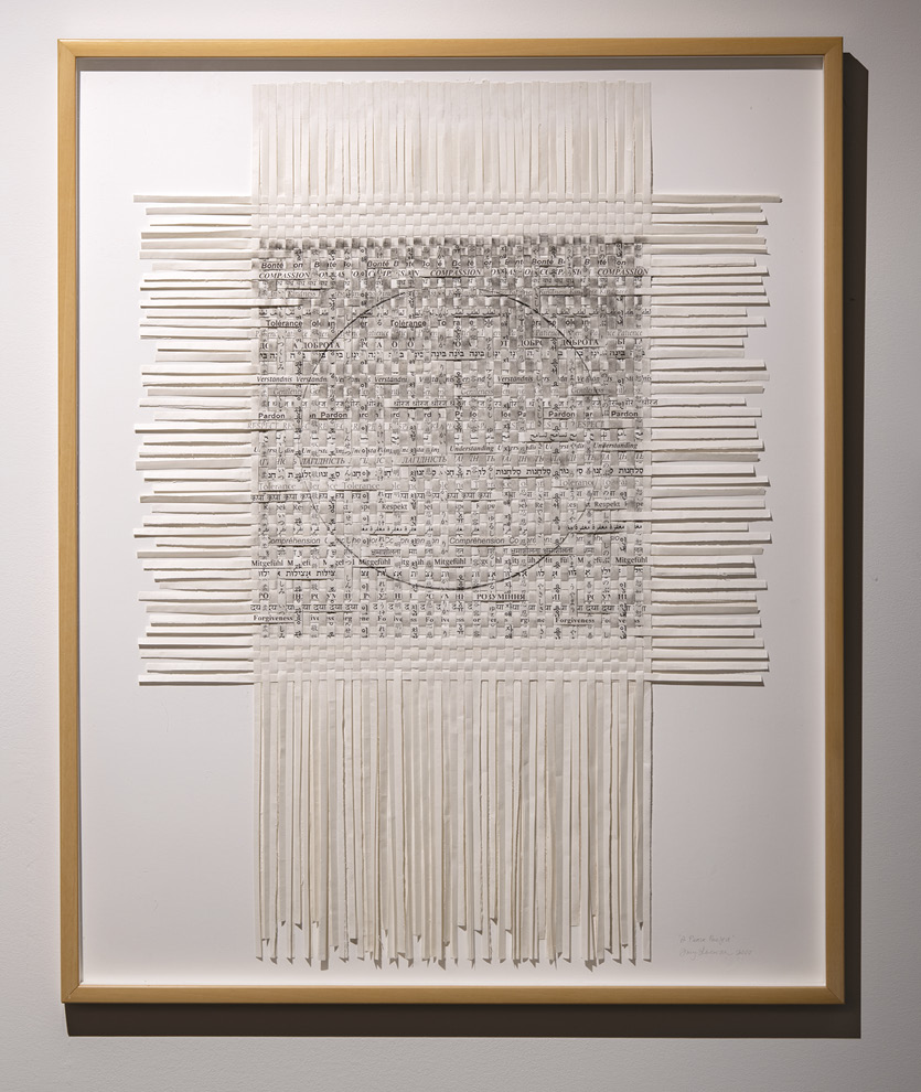 Photo: a mounted and framed series of vertical and horizontal strips of white material woven in the middle with some visible words in different languages — forgiveness, compassion — and a black circle overlaid in the middle.