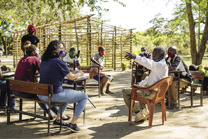 Colour photograph: Photo of about 9 people sitting outside in a circle on benches and plastic chairs. A tree and framing for a building are in the background.  An elderly man gesticulates as he talks.
