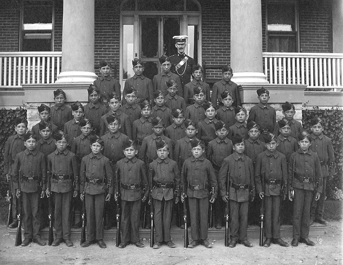 Black and white photograph: Five rows of male Indigenous students of various ages formally posed in cadet uniforms on the steps of the Mohawk Institute.  Their male teacher, also in uniform, stands behind them. The students are standing, with firearms at their side.