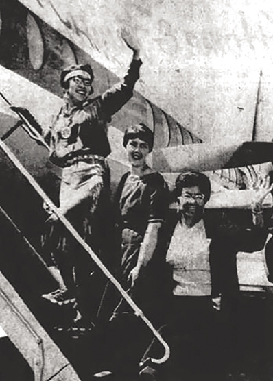 Black and white photograph of three women going up the stairs to a plane. The first woman wears a headband and glass and looks backward, waving.