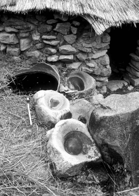 Three broken grindstone-mortars found in front of a stone house with a thatched roof.