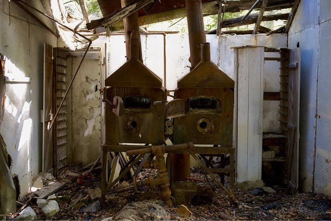 Two gold smelter structures sit in a long abandoned building. Light is streaming in collapsed doors and in ceiling gaps of the building to illuminate the once productive smelting structure.