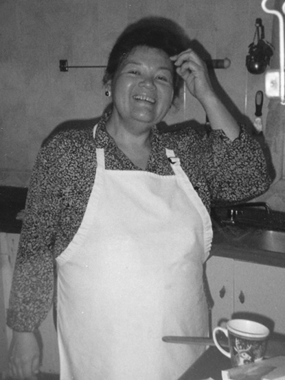 Black and white photograph: Adult Indigenous woman in a kitchen wearing a light-coloured apron. There is a cup in front of her on a table and she is touching her forehead.

