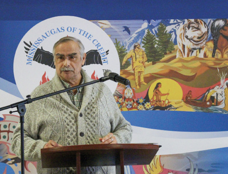 Colour photograph. An Indigenous man is standing at a lectern speaking into a microphone. He has short grey hair and a moustache, and is wearing an olive knitted cardigan. The wall behind him has the round Mississaugas of the Credit logo, showing an eagle in flight and fires, and there is a mural with First Nations people, wolves, canoes, trees, and mountains.