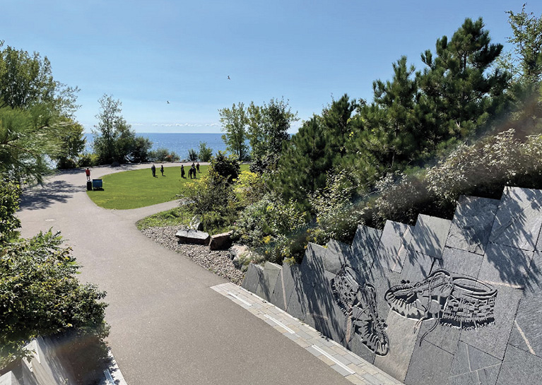 Colour photograph. A park on a lakeshore, with a large footpath, many large trees, and a retaining wall on the right made of stone slabs. There are images of two large moccasins carved into the wall. A group of people are walking along the grass towards the lakeshore.