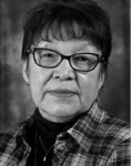 Black and white photograph: Portrait of a woman with short hair, glasses, and wearing a collared plaid shirt over a turtleneck with delicate necklaces.