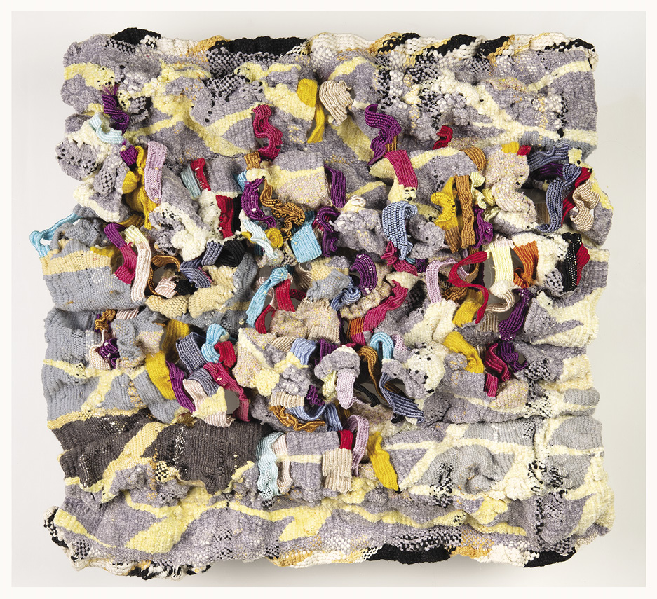 Photo: a large multi-colour jumble of textiles. It is mostly grey with yellow stripes with blue, red, mustard yellow, purple ribbons of cloth coming out of it. 