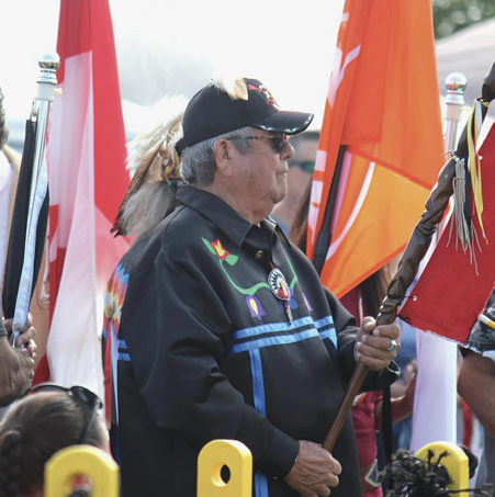 Colour photograph. A man standing holding a carved staff decorated with coloured ribbons He is wearing a black ribbon shirt with floral beadwork on the front, a black baseball cap, and sunglasses. There are several flags behind him, and a crowd is visible around him.