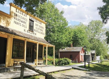 Colour Photograph. Historical recreation of a nineteenth century streetscape. A yellow general store is in the foreground, while a pink clapboard building is in the background. Many green trees are in the area.