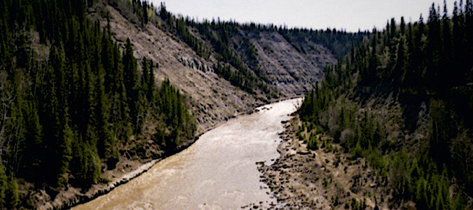 In northeastern British Columbia near Dawson Creek, the fast flowing Kiskatinaw river carves through banks of steep evergreen tree forested mountains of a dark green with brown rock slopes. The flow of the water has felled trees and loosened the rocks to wash out wider banks with a growing rock shore.