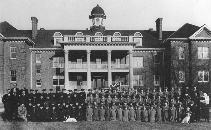 Black and white photograph: Outside view of the front of the school building, which is a large, wide, brick building with three storeys, with sizeable pillars holding up the multi-storey front porch. Approximately eighty Indigenous students ranging in age are formally posed in rows in front of the school. On the left are boys in a dark uniform and on the right are female students, also in a uniform that is a bit lighter in colour.  Teachers are also in the photograph.