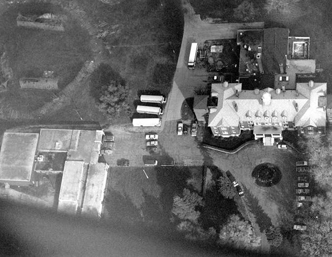 Black and white photograph: Aerial photograph of the third Mohawk Institute.  There is a large school building on the right with numerous smaller buildings behind attached to the school.  To the left are several outbuildings and grassed areas.  There is a large circular drive located in front of the school, and school buses and a variety of vehicles are parked around the school.  The image appears to be approximately 1960s or 1970s.