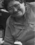 Black and white photograph: Photograph of a woman wearing a collared shirt and glasses, leaning in to be in the photograph. It appears to have been cropped from a larger photograph.
