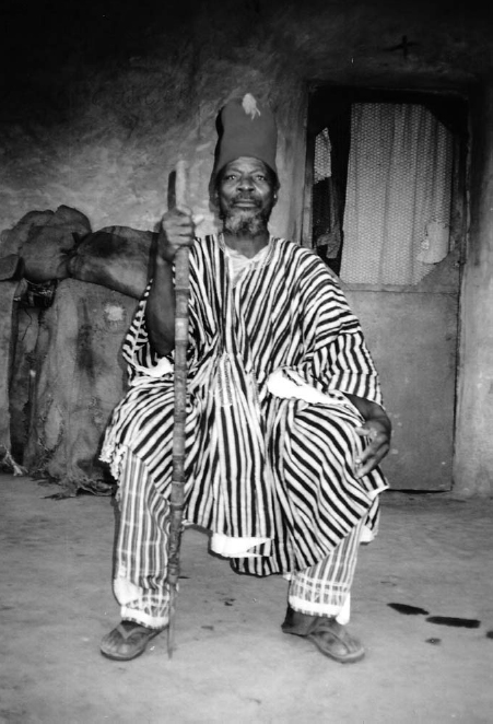 A man in striped traditional attire carries a stick and sits inside a stone room. A door and a pile of sacks are seen behind him.