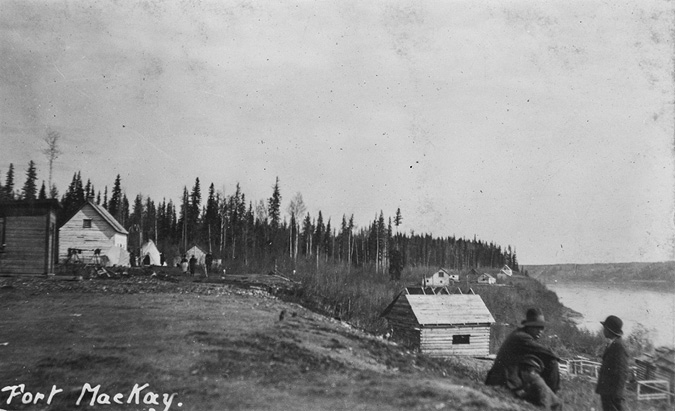 Black and white photograph:a small settlement area along a riverbank, identified as Fort McKay. In the foreground, two men wearing hats are seated on an uneven grassy terrain, looking towards the buildings and river. To the left, several wooden structures with peaked roofs are visible, including a couple of tents. These buildings are situated close to a dense forest of tall, slender coniferous trees. The Athabasca river runs parallel to the settlement, framed by the wooded landscape. The sky appears overcast, lending a subdued atmosphere to the scene.