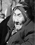 Black and white photograph: Portrait of a woman in profile, sitting in the bright sun, wearing a winter wool coat and a wrapped head covering. 