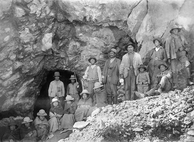 A mine-workers’ team pose at the entrance of a mine. Men are standing and women are sitting. There is a young boy next to an older man and close to the only sitting young man. While men wear Western styled attires, women are dressed with native garments.
