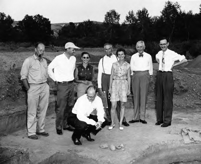 A photograph shows several people inspecting a piece of land, Trees and mountains can be seen in the background.