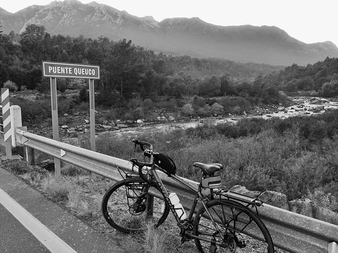Black and white photograph: Photo of a bicycle parked near a “Queuco Bridge” sign, with the river running below and mountains in the background