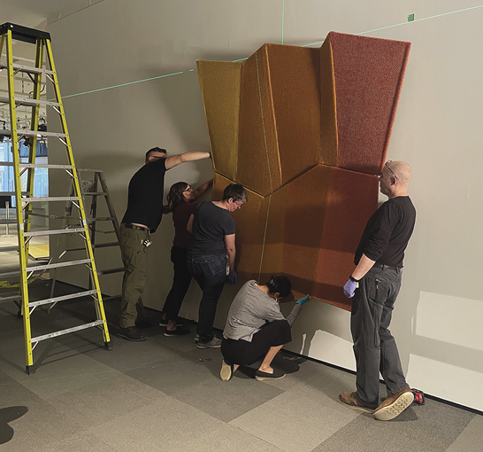 Photo: five people working to hang a large section of art on a white wall. A yellow ladder sits to the left.