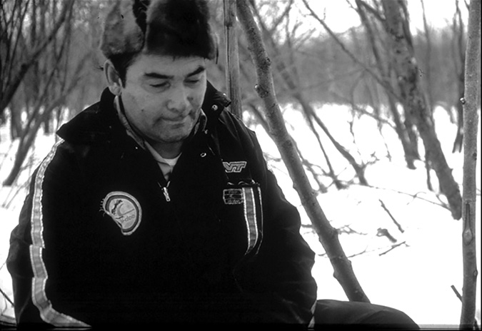 The black-and-white image shows a man sitting outdoors in a snowy, wooded area. He is wearing a dark jacket with a fur-trimmed hood and noticeable emblem patches. His gaze is directed downward, and his expression appears contemplative. Bare trees surround him, their branches reaching skyward, creating a textured backdrop against the blanket of snow on the ground. The snow contrasts starkly with the dark tones of his clothing and the trees.
