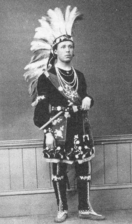 Black and white historical photograph. A young man is standing wearing Indigenous regalia. He is wearing a large, feathered headdress, a dark tunic with floral designs at the bottom, an elaborate sash and bandolier bag, and decorated leggings and moccasins. He has a tomahawk tucked into his belt.