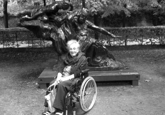 Black and white photograph. An older woman sits in a wheelchair, outside, in front of an abstract bronze statue. Her hands are folded, and she is holding a white handbag. 
