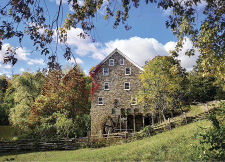 Colour photograph. A tall stone mill with several storeys stands behind a fence, surrounded by mature trees. There is a dimly visible pond to the left. The mill has eight windows on the visible wall, and some vines in autumn colours along it.