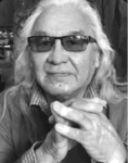 Black and white photograph: Portrait of a man wearing tinted glasses and a collared shirt, his hand held up in front of his chest with fingers interlaced.