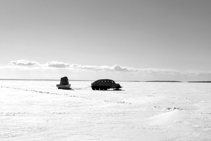 Black and white photograph, a frozen lake stretches into the horizon on a clear day; a snowmobile pulls a canoe on sleds, a large squared camera obscura fills the canoe.