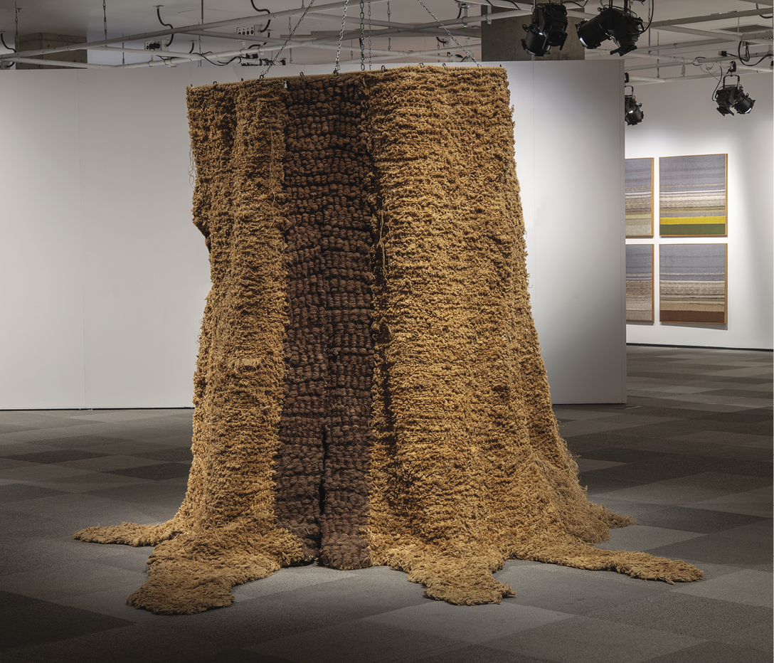 Photo: large tree trunk shaped weaving in a gallery setting. It is mostly brown with a darked strip in the middle.