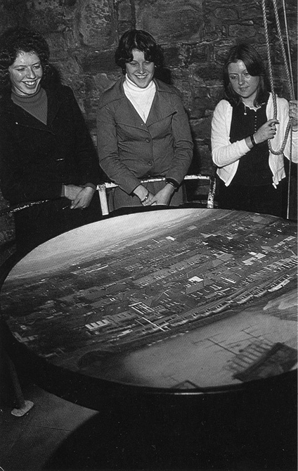 Black and white photograph, three young women stand smiling looking at a large circular dish inside a camera obscura, showing a port with dense terraced houses.