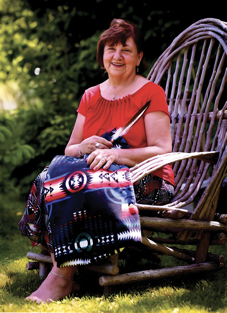 Colour photograph. Portrait of an Indigenous woman seated on a large wooden chair. She has mid-length brown hair and is smiling. She wears a short-sleeved red blouse, and has a First Nations blanket on her lap. She is holding an eagle feather in her right hand.