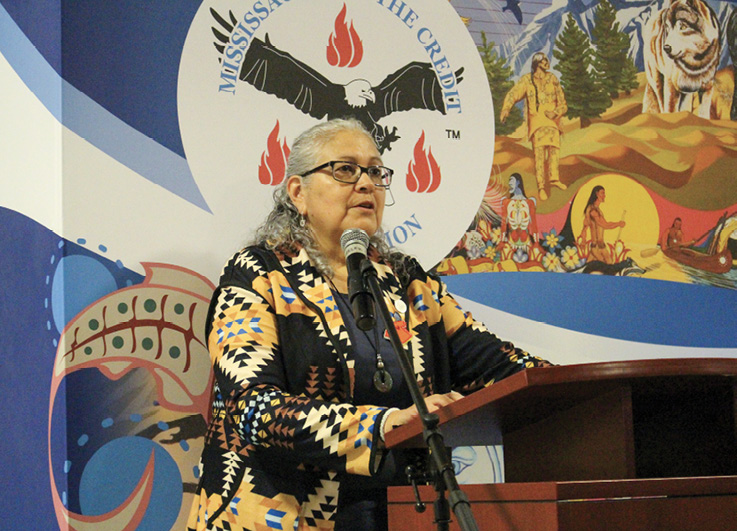 Colour photograph. An Indigenous woman is standing at a lectern speaking into a microphone. She wears glasses, and has long grey hair tied back at the temples but hanging to her shoulders . She is wearing a black and yellow jacket with First Nations arrow designs, and a pendant necklace. The wall behind her is decorated with the round Mississaugas of the Credit logo, showing an eagle in flight and fires, and beside it there is a mural with First Nations people, wolves, canoes, trees, fish, and mountains.