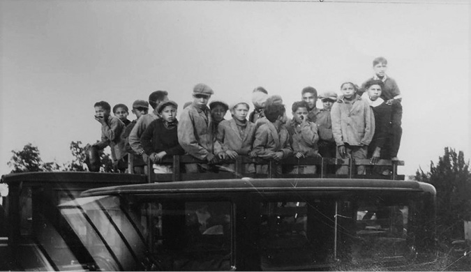 Black and white photograph: Group of approximately twenty Indigenous male students of the Mohawk Institute standing in the uncovered bed of a large truck.  Most are wearing coats and a cap.  They appear to be crowded into the truck bed.