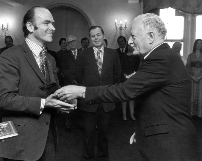Black and white photograph: A man in a suit and tie, Clive Baxter, in the left of the photo receives a trophy from another man in a suit on the right, His Excellency Roland Michener. Michener is speaking to Baxter. A group of well dressed people watch on in the background.