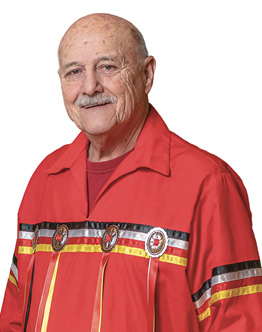 Colour photograph. Head and shoulders portrait of a man, smiling. He is bald and has a grey moustache. He is wearing a red ribbon shirt with black, white, red and yellow silk ribbons running horizontally along the front of the shirt, There are three medallions on this band of four horizontal ribbons, with red and yellow ribbons running down from them.