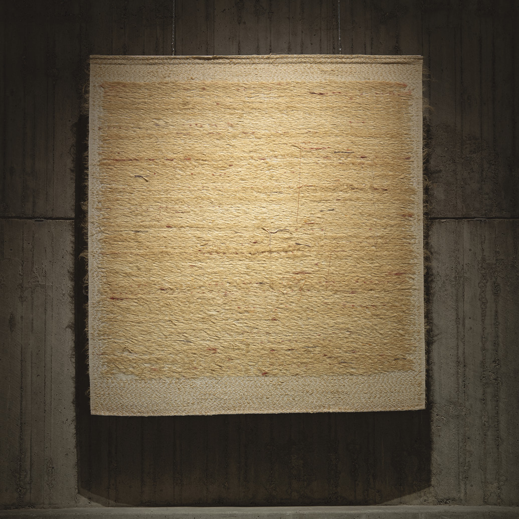 Photo: spotlit hanging of an entirely tan-coloured weaving composed of many horizontal lines. Sections look rough with strings poking out the sides of the piece.