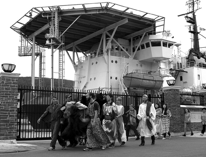Black and white photograph, a large group of people wearing a variety of togas, dresses, and modern jeans and sneakers push a taxidermized moose on wheels, a large ship looms over a fence behind them.
